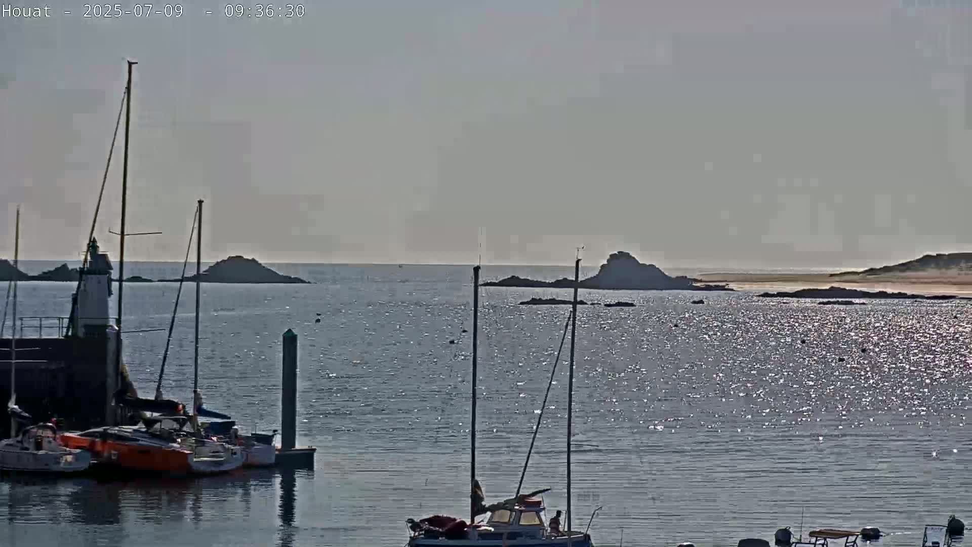 Several sailboats are moored near a lighthouse in a calm harbor, with rocky islands and a sandy beach visible in the distance under a hazy sky.
