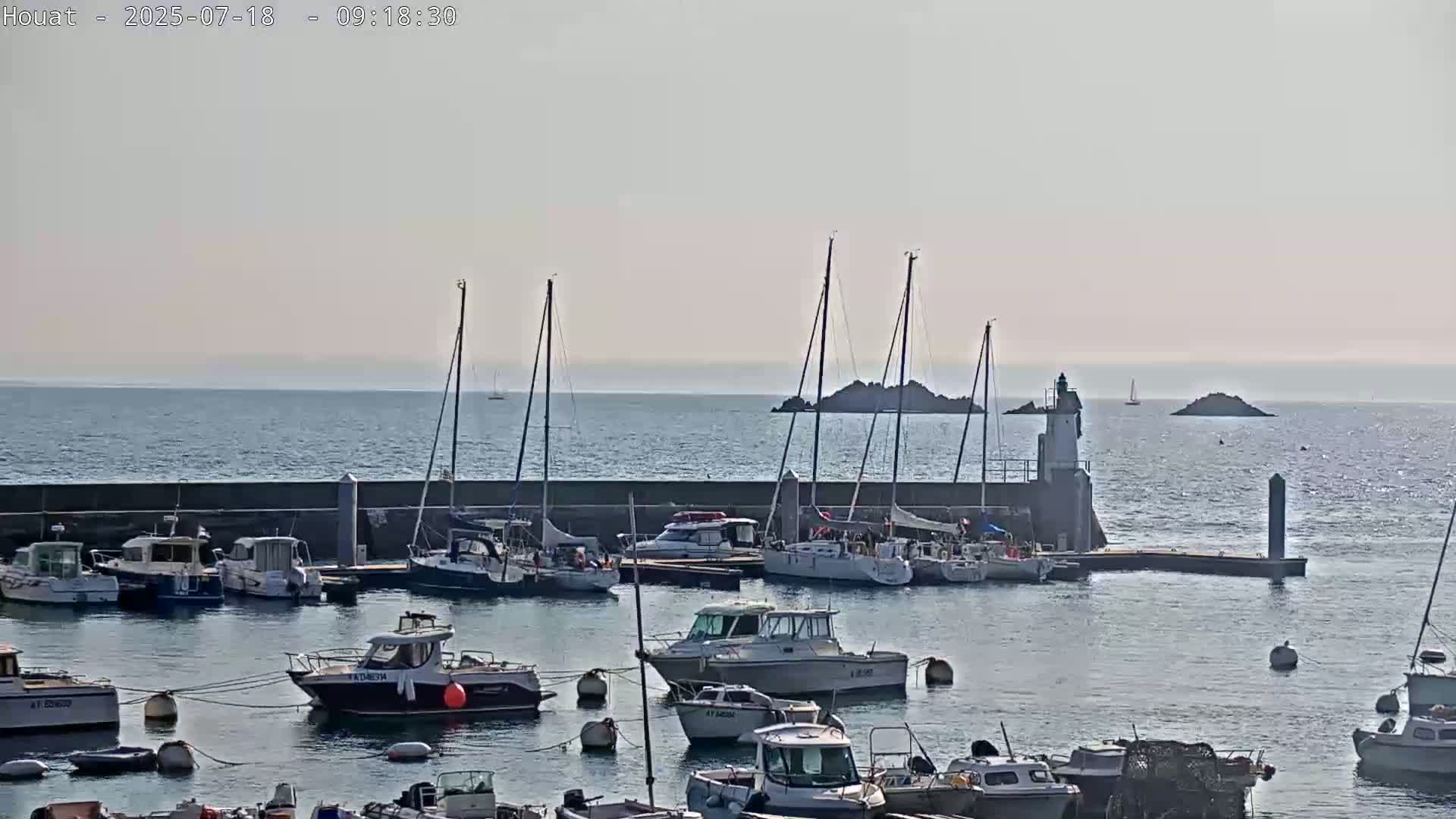 A calm, sunny day shows a harbor filled with numerous boats of varying sizes, moored alongside a low stone wall, with a lighthouse and small islands visible in the distance.