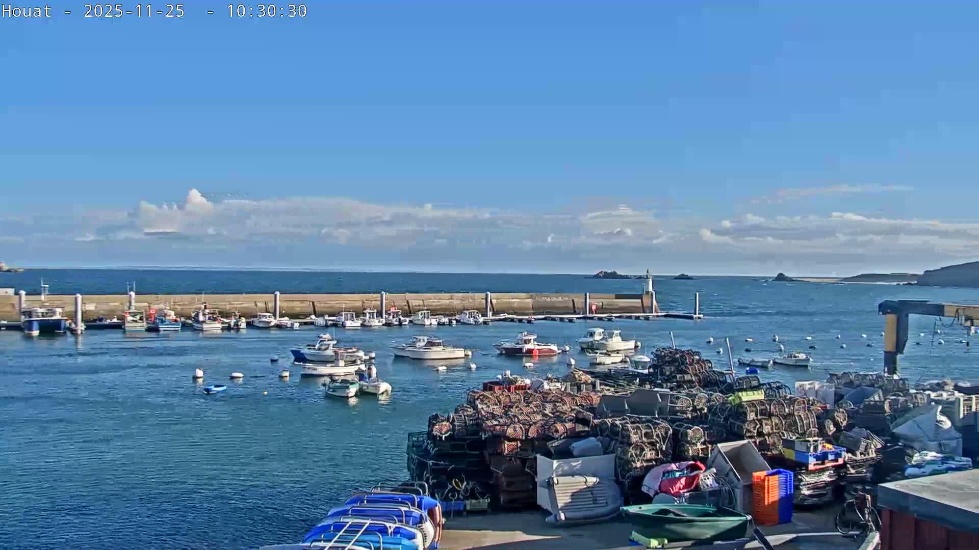A sunny day at a bustling harbor features numerous boats moored in the clear blue water behind a concrete breakwater, a small lighthouse, and large stacks of lobster pots and other fishing gear on the dock in the foreground, all under a mostly clear blue sky with scattered white clouds.