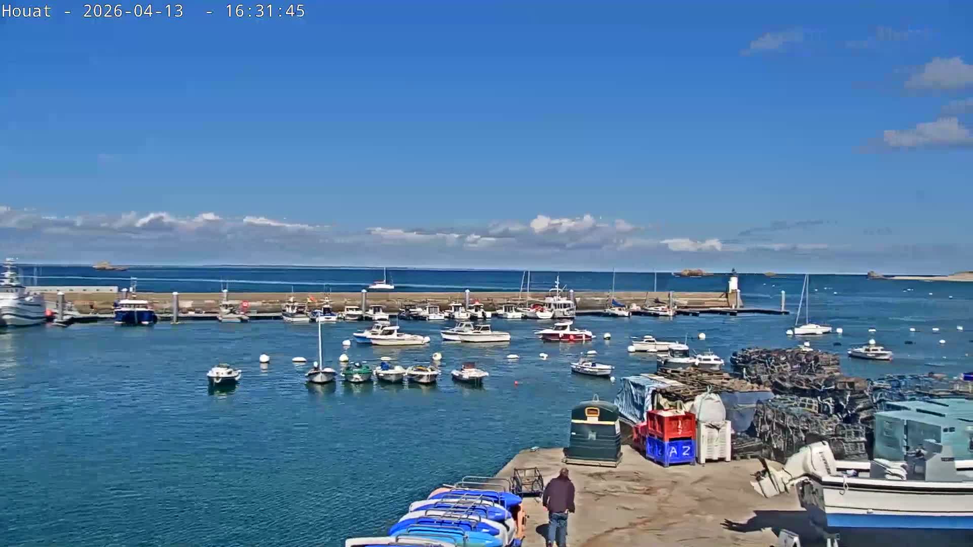 A sunny day at a bustling harbor features numerous boats moored in the clear blue water behind a concrete breakwater, a small lighthouse, and large stacks of lobster pots and other fishing gear on the dock in the foreground, all under a mostly clear blue sky with scattered white clouds.