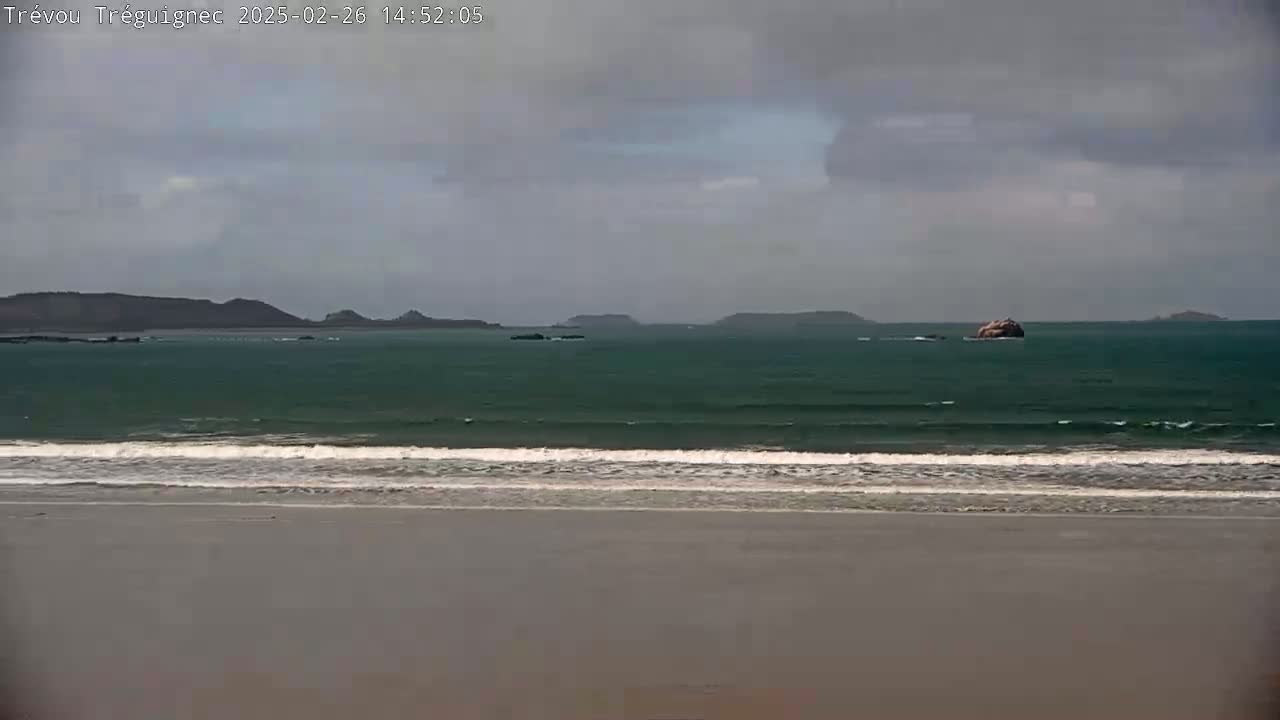A sandy beach stretches to a dark green ocean with small waves under a cloudy sky, and distant islands are visible on the horizon.