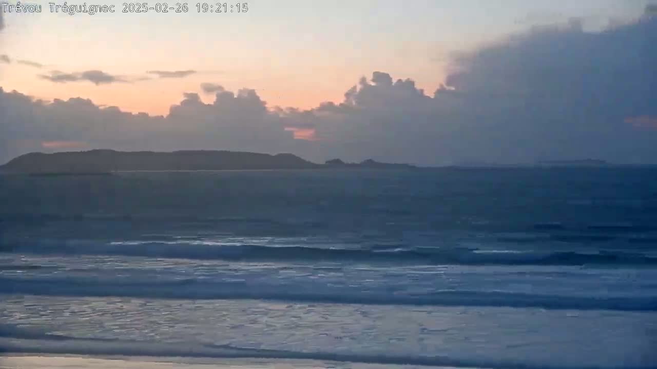A mostly cloudy sunset over a calm ocean with gentle waves rolling onto a sandy beach.