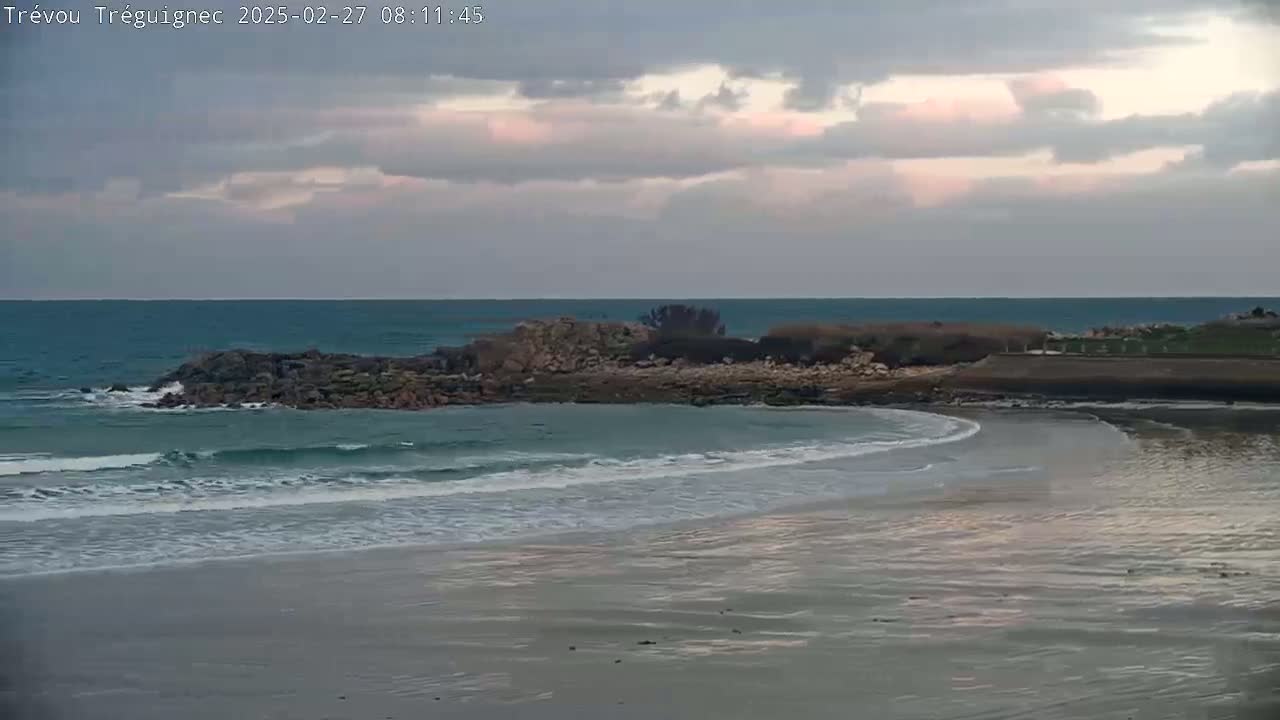 Under a cloudy sky, small waves lap a sandy beach near a rocky outcrop that juts into a calm ocean.