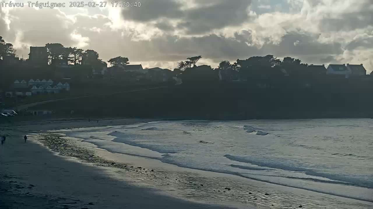 A sandy beach with gentle waves under a cloudy sky overlooks a hillside with houses and trees.