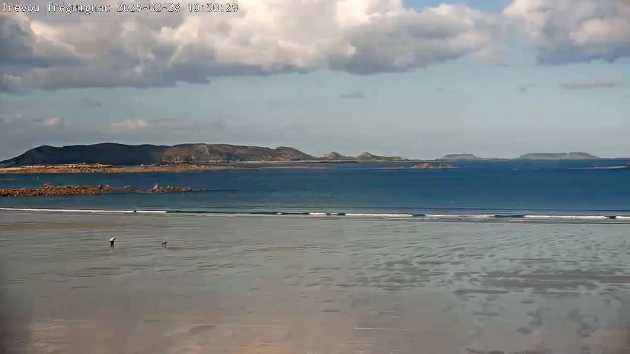 A mostly cloudy day reveals a wide sandy beach with small waves lapping the shore, a distant rocky shoreline, and two people walking on the wet sand.