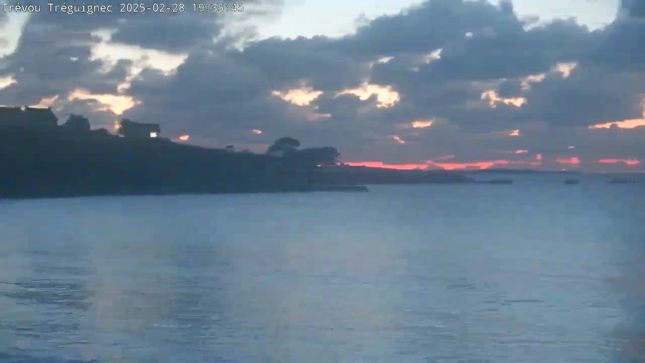 A calm body of water reflects a sunset sky filled with mostly dark clouds, with a sliver of pink and orange visible on the horizon.