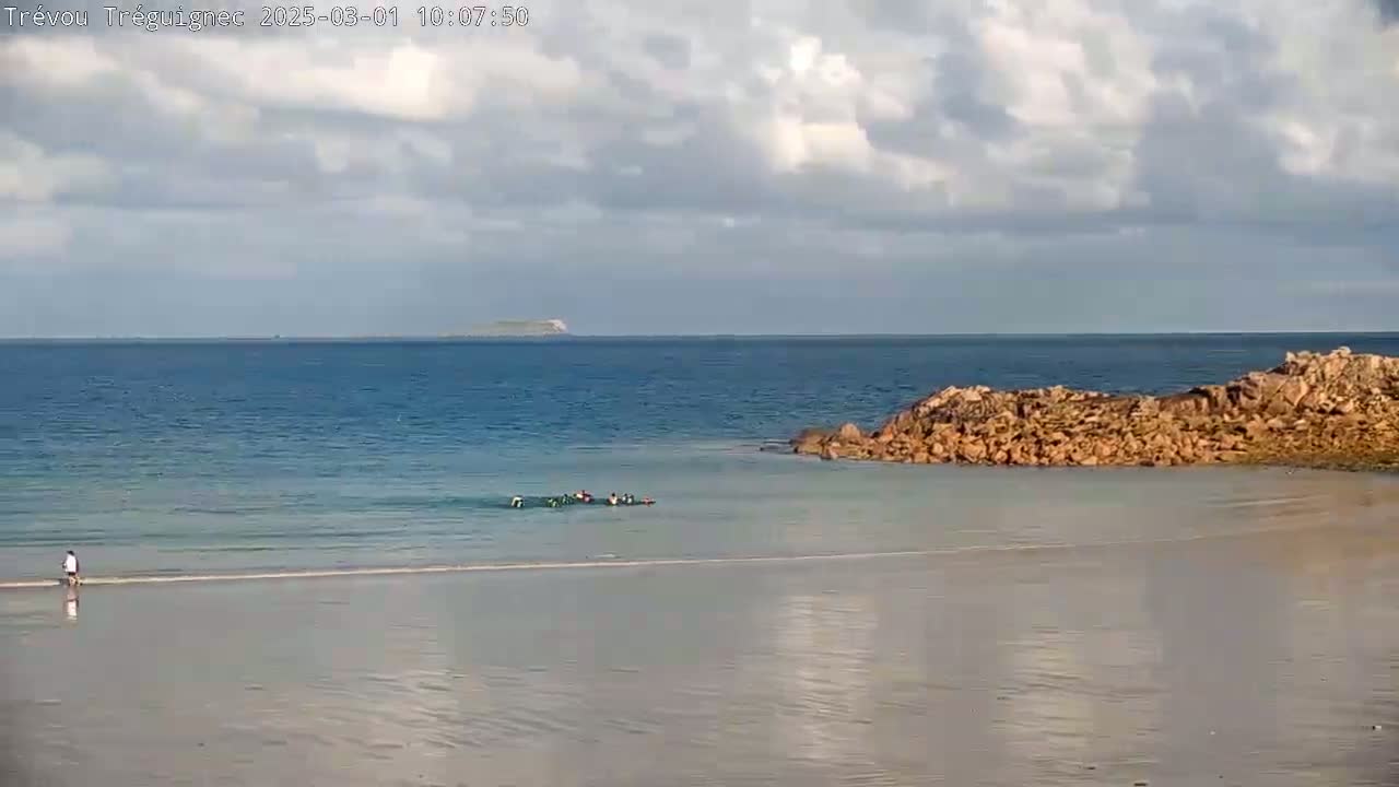 A mostly cloudy day shows a sandy beach, calm ocean water, a rocky outcropping, a group of people in the water, and a distant island.
