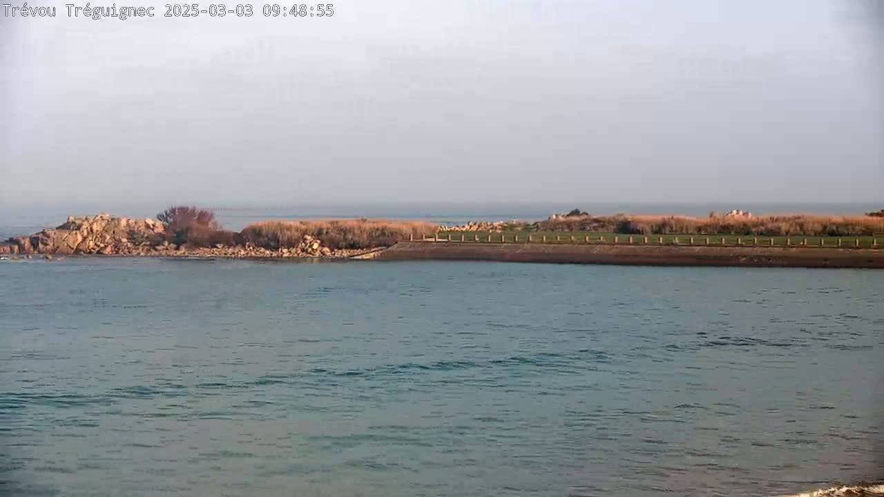 A rocky outcrop and grassy area are visible across a calm body of water under a hazy sky.