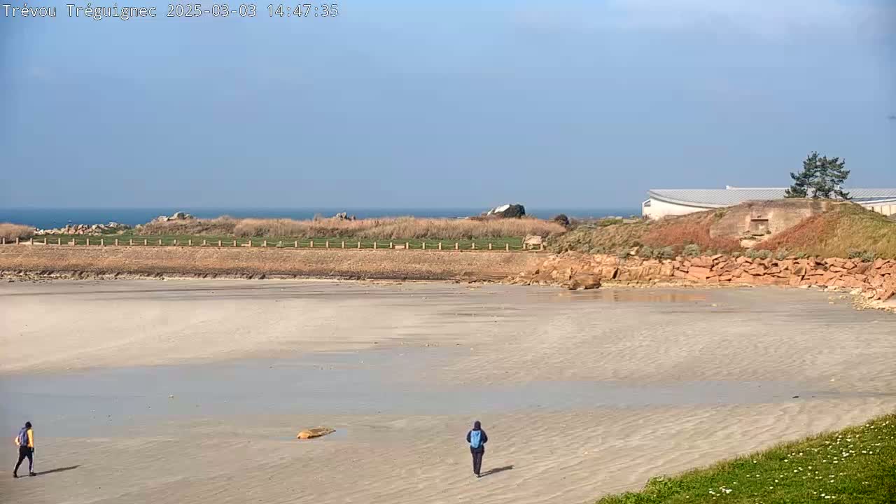 Two people walk on a wide, light-colored sandy beach under a clear blue sky, with a calm ocean in the distance and a building on a rocky outcrop.