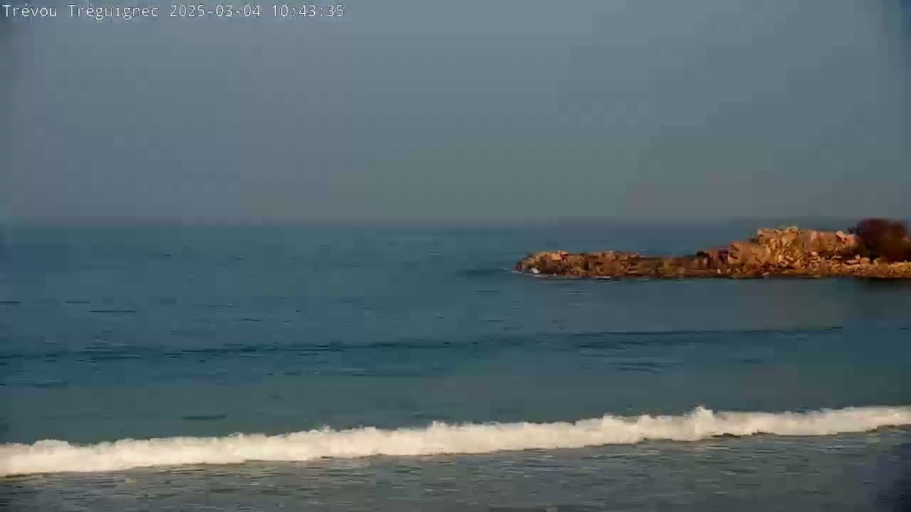 A rocky outcrop sits in calm, teal ocean water with small waves breaking on the shore under a hazy sky.