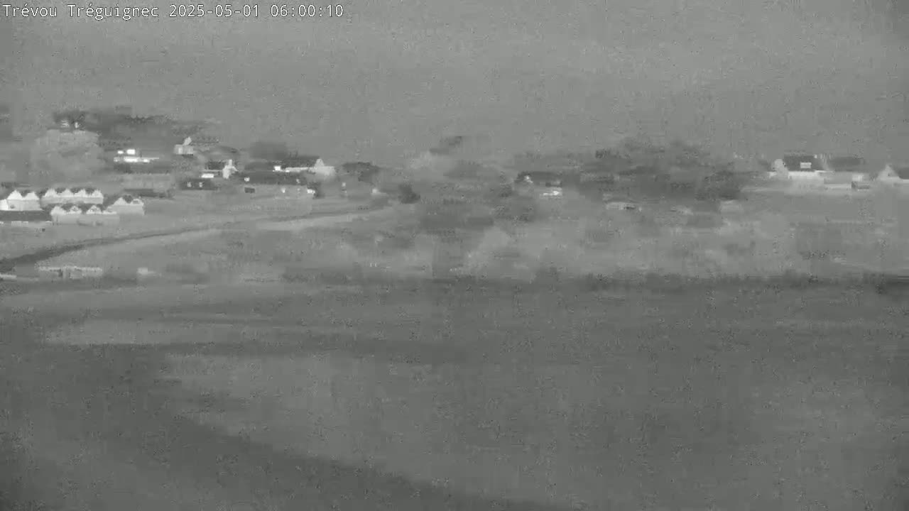 A grayscale thermal image shows a small village at dawn or dusk, with low-lying clouds or fog obscuring the sky.