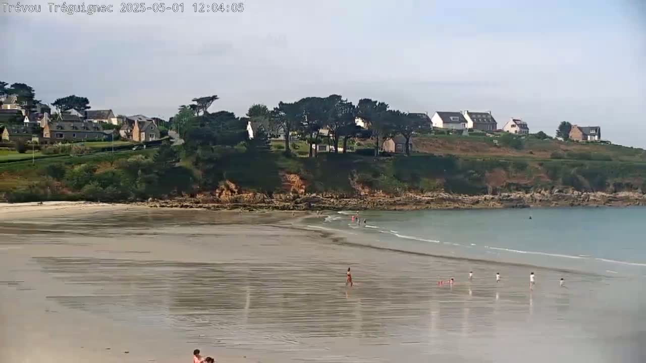 A mostly sunny day at a sandy beach with calm, light blue water, several people wading in the shallows, and a small village on a hillside overlooking the beach.