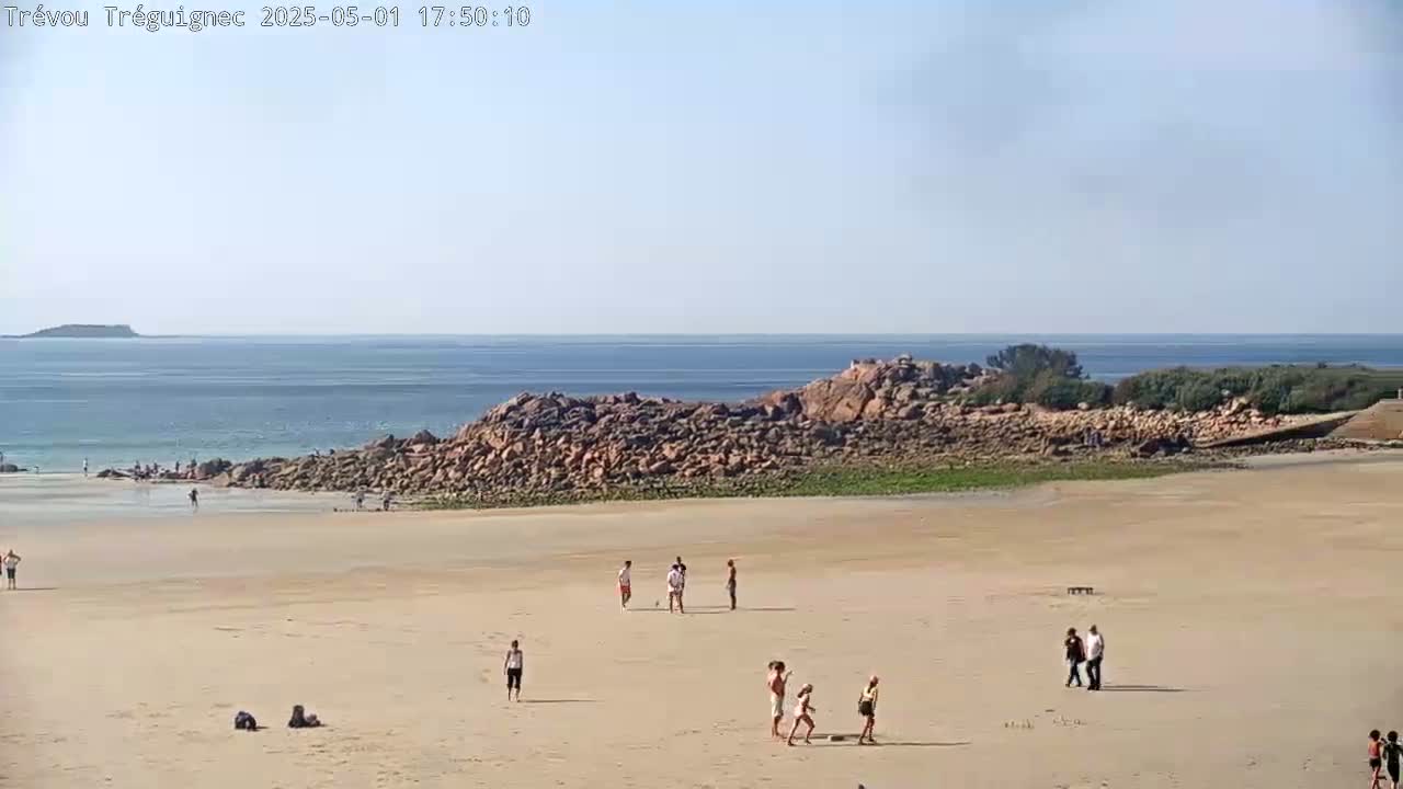 A sandy beach with a few people scattered across it, backed by a rocky outcrop and a calm ocean under a clear sky.