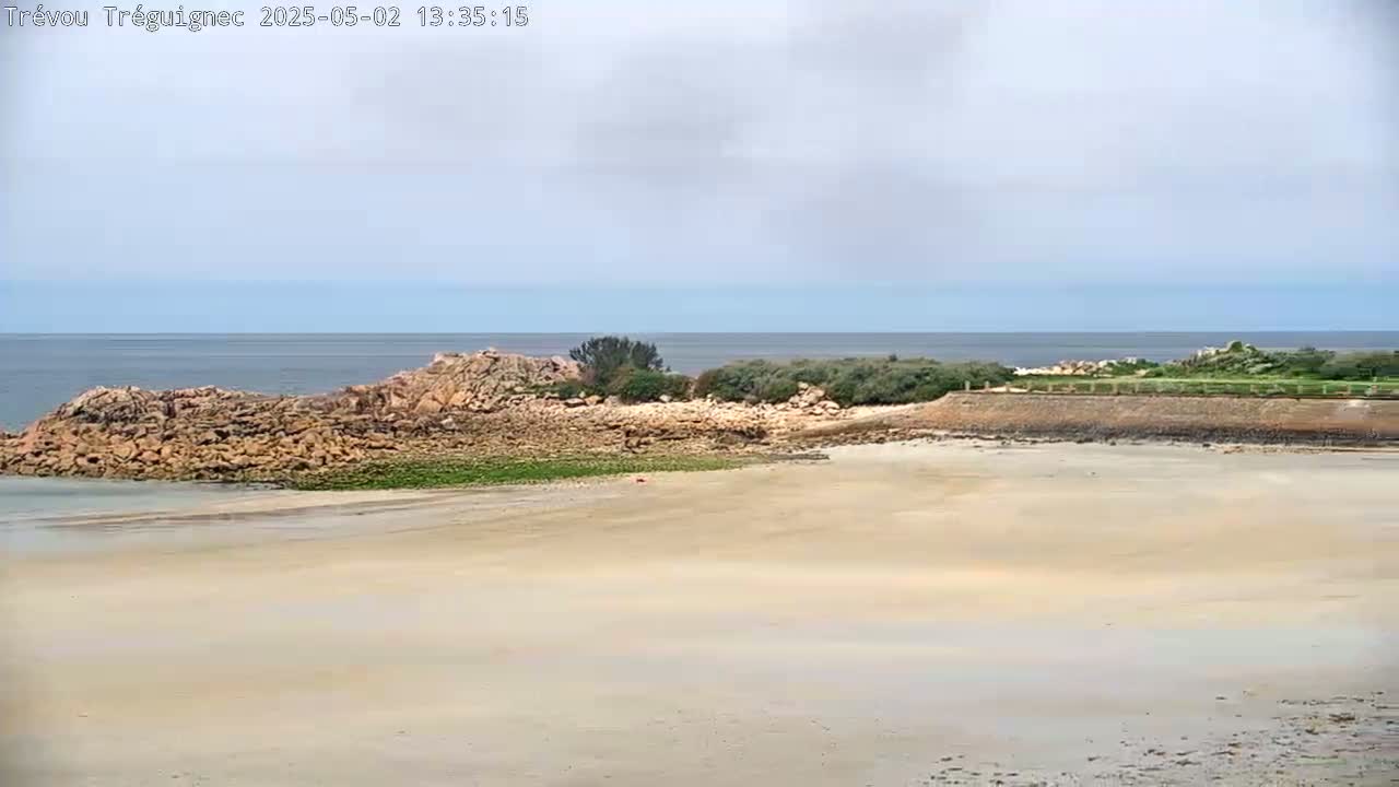 A sandy beach stretches out to a rocky outcrop separating it from a calm ocean under a mostly cloudy sky.