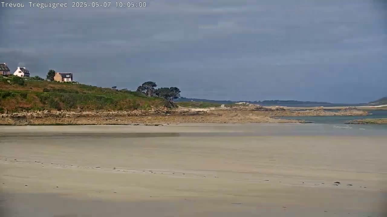 A sandy beach stretches to a rocky shoreline, beyond which lies calm water and a low-lying land with a few houses under a mostly cloudy sky.