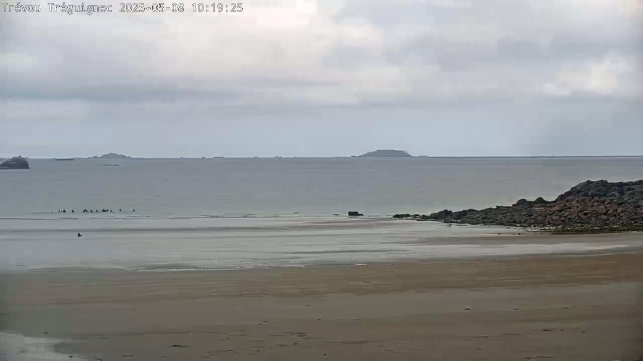 A sandy beach stretches to a calm, gray sea under a cloudy sky, with distant islands visible on the horizon.