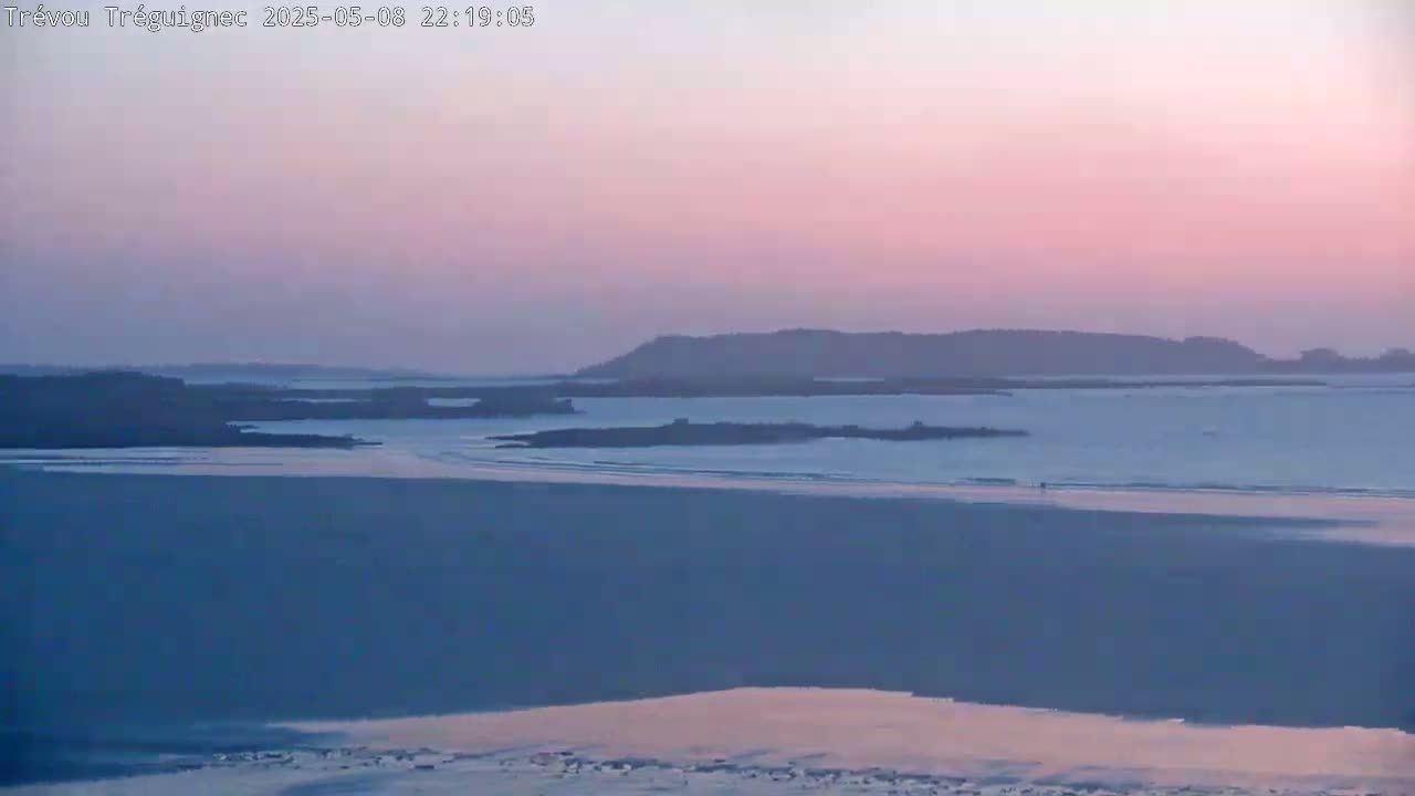 A pink and purple sunset over a calm body of water, with dark silhouettes of land in the distance and a flat, sandy beach in the foreground.