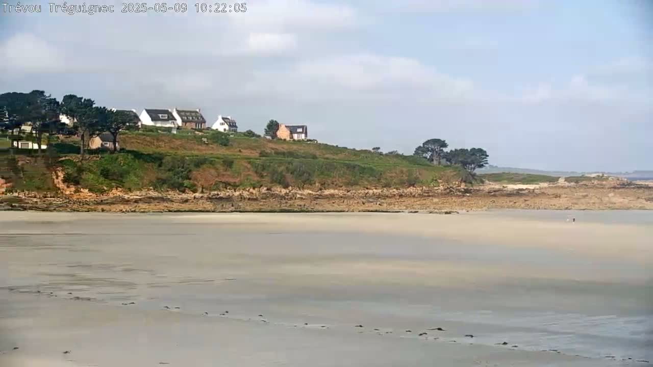 A wide sandy beach stretches out to a rocky shoreline where several houses sit atop a grassy hill under a partly cloudy sky.