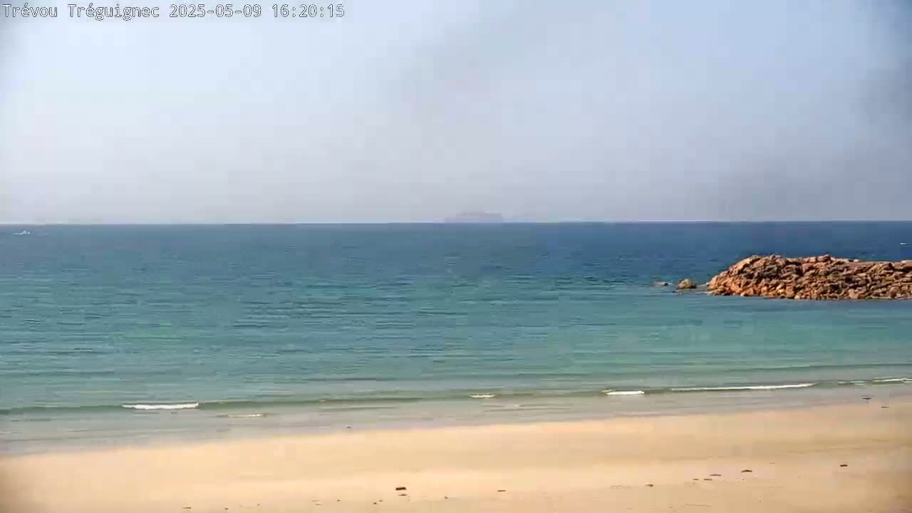 A sandy beach meets a calm, blue ocean under a hazy sky, with a rocky outcrop in the water.