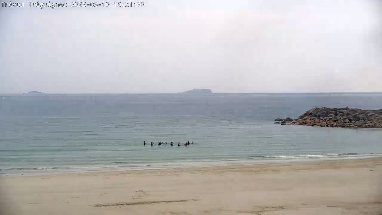 A sandy beach and calm, light-blue ocean with a few people in the water and a small rocky outcrop near the shore, under an overcast sky.