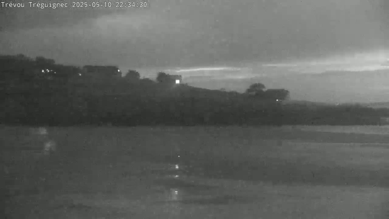 A nighttime, overcast scene shows a dark hill with a few structures and some trees, overlooking a calm body of water.