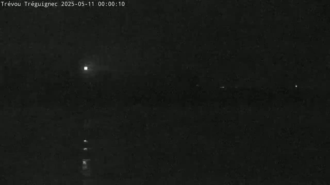 A nighttime, dark image shows a body of water reflecting a bright light source in the distance under a mostly clear sky.