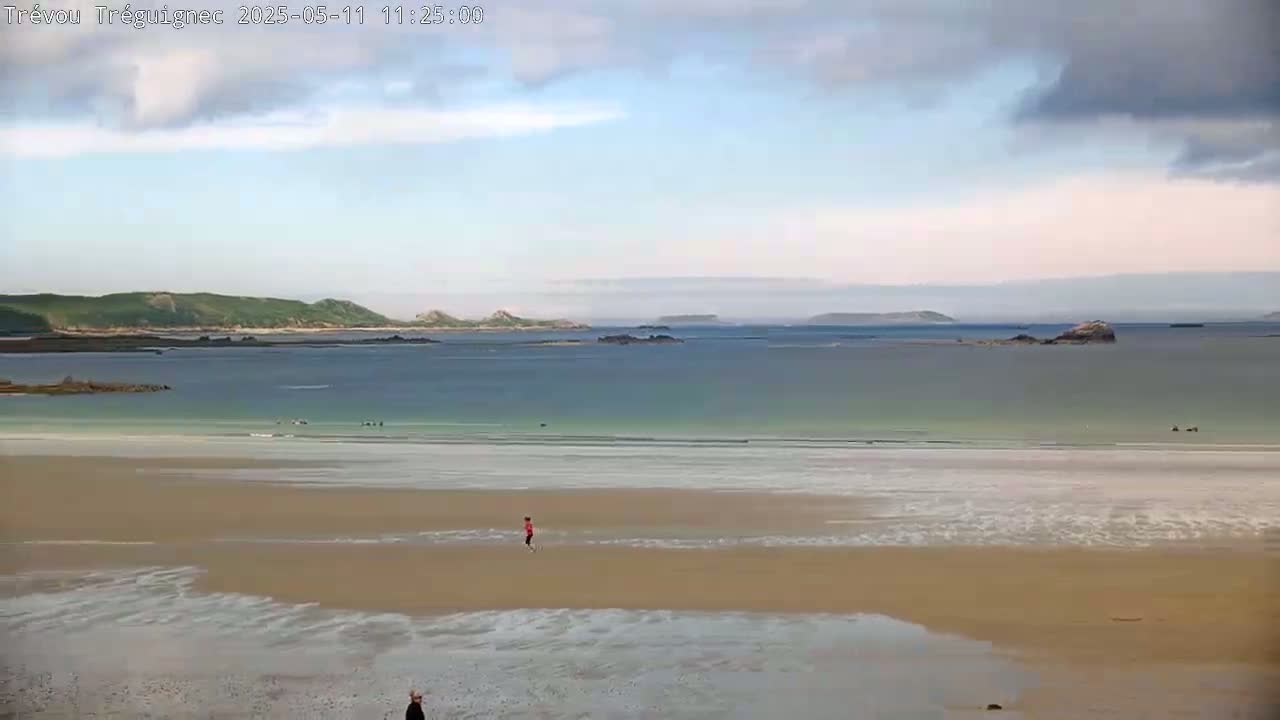 A person walks on a wide, sandy beach, with calm, light-green water stretching to distant islands under a partly cloudy sky.