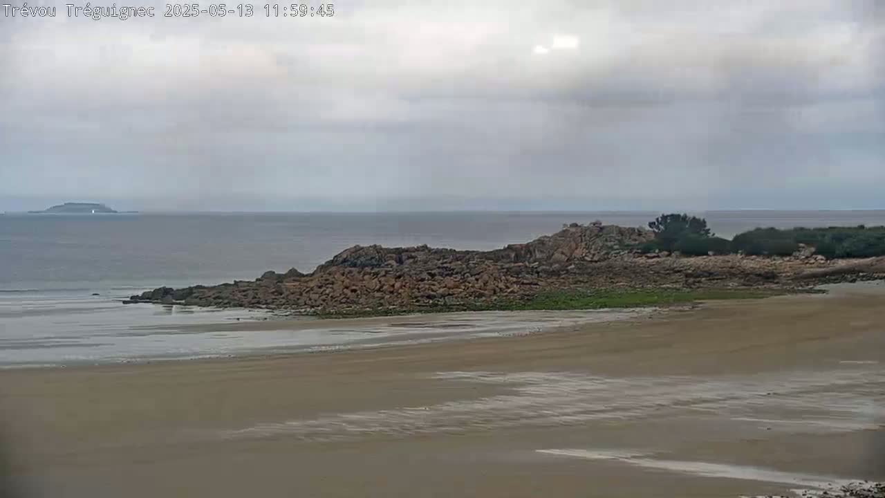 A sandy beach stretches out to a rocky outcrop, behind which lies a calm sea under an overcast sky.