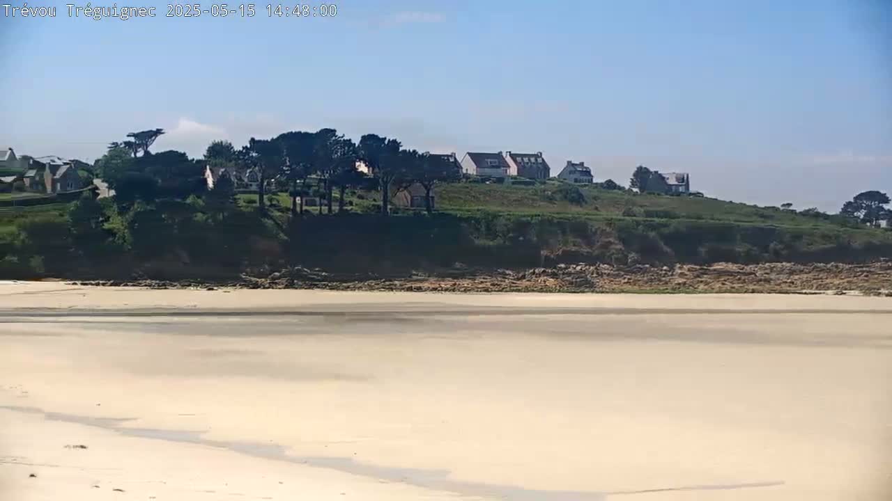 A sandy beach stretches out to a grassy hill with houses and trees under a clear, sunny sky.