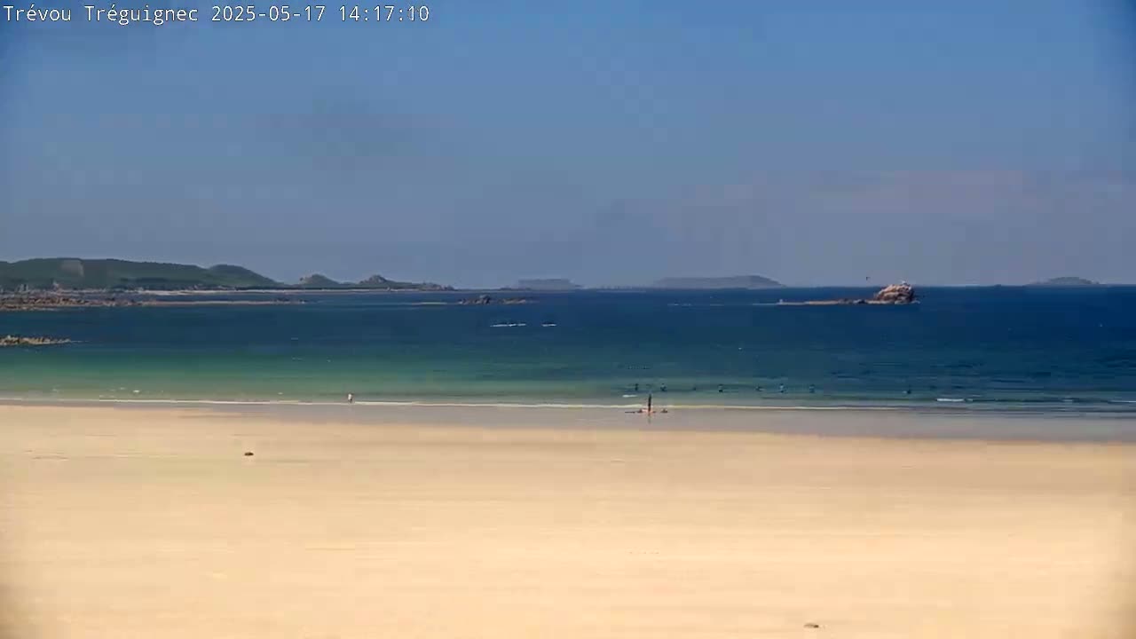 A wide sandy beach stretches toward a calm ocean under a clear blue sky, with a few people visible in the water and on the sand.