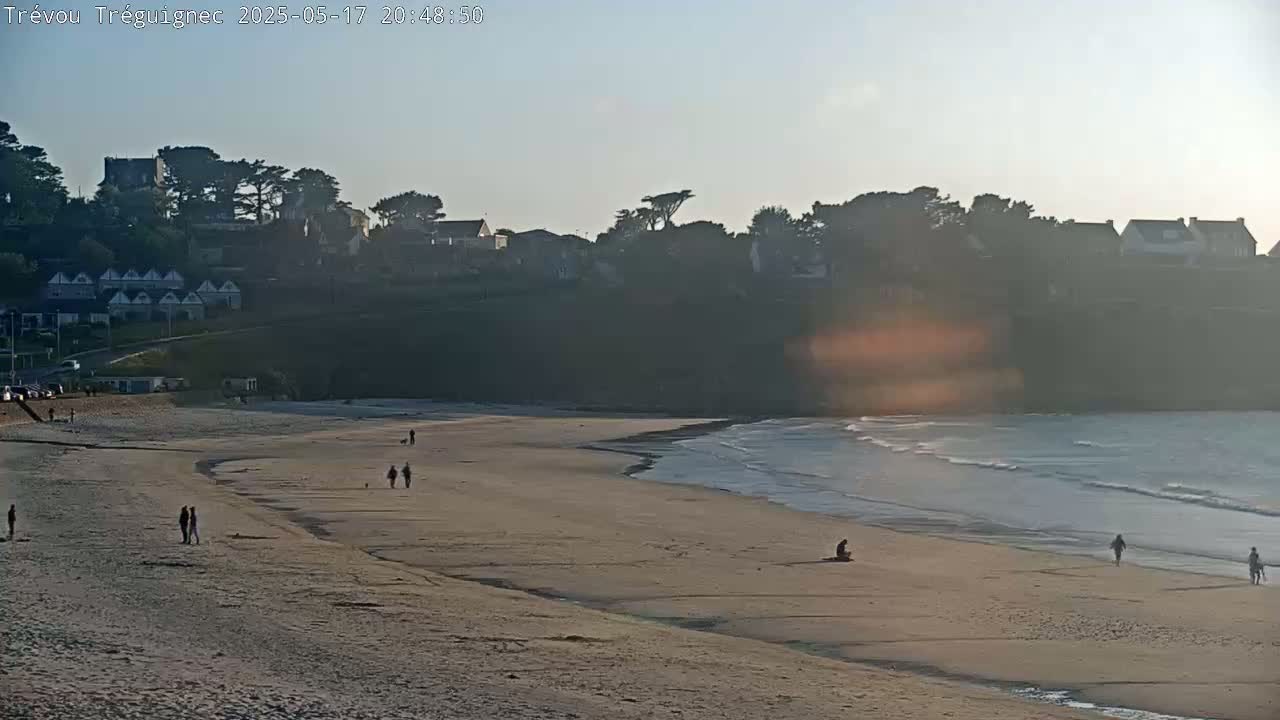 A sandy beach with a few people walking on it, overlooks a calm ocean under a clear, bright sky.