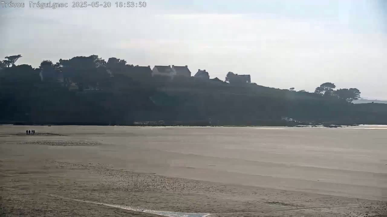 A wide sandy beach stretches out to a hazy, dark hilltop with a few buildings and trees under a mostly overcast sky.