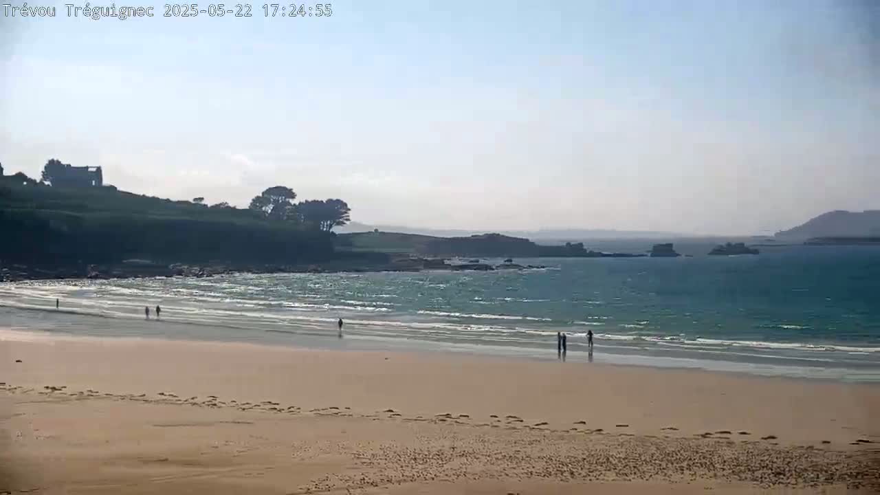 A sandy beach with a few people on it stretches out to a calm, light-blue ocean under a hazy sky.