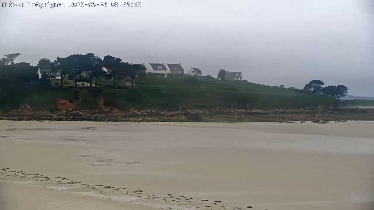 A sandy beach stretches out to a rocky shoreline, behind which sits a cluster of houses on a hill under an overcast sky.