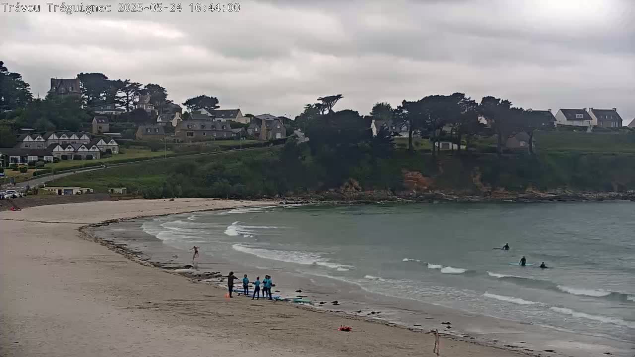 A sandy beach with several people standing near surfboards, some surfers in the water, and a village on a hill overlooking a calm, grey ocean under an overcast sky.