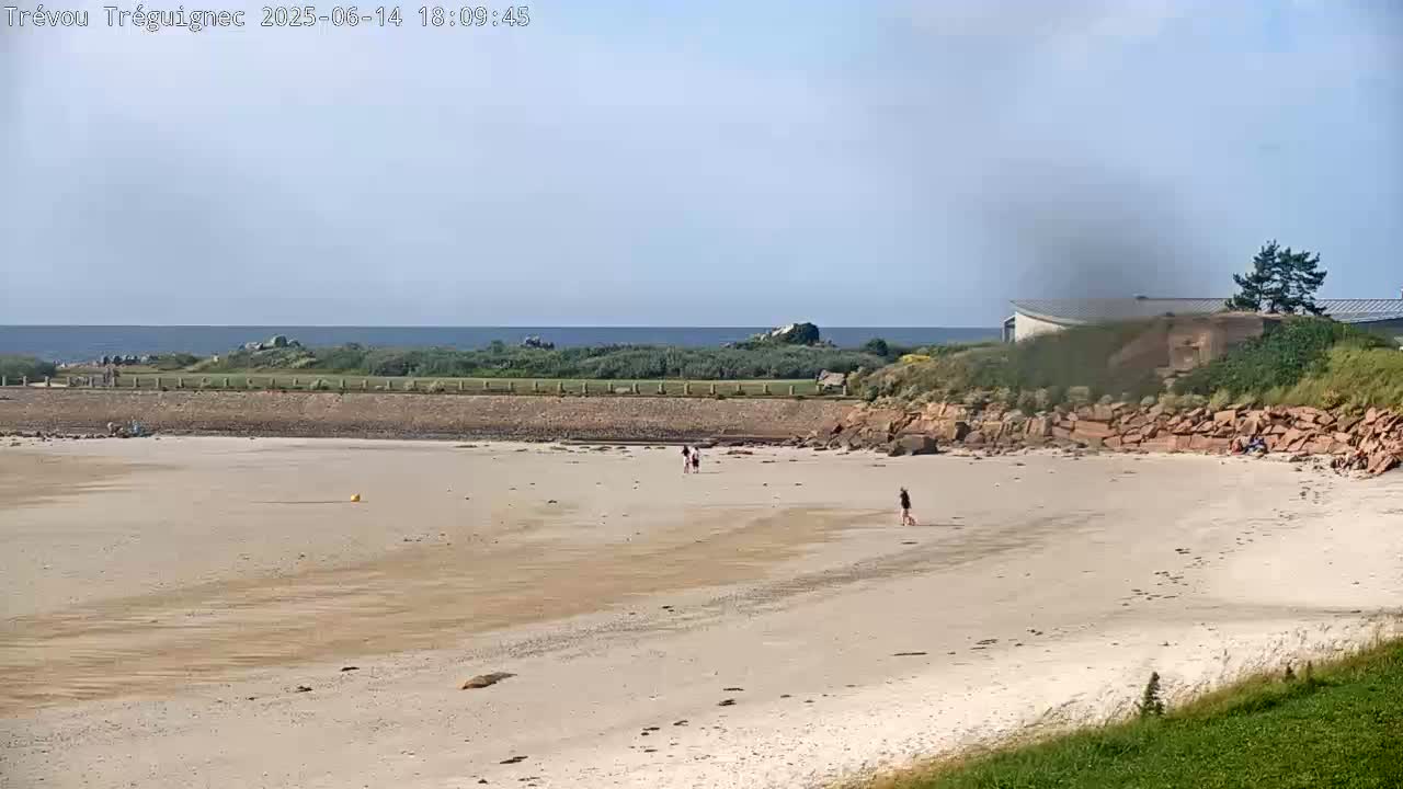A mostly empty sandy beach stretches toward a calm ocean under a partly cloudy sky, with a few people walking on the sand and a building visible in the distance.