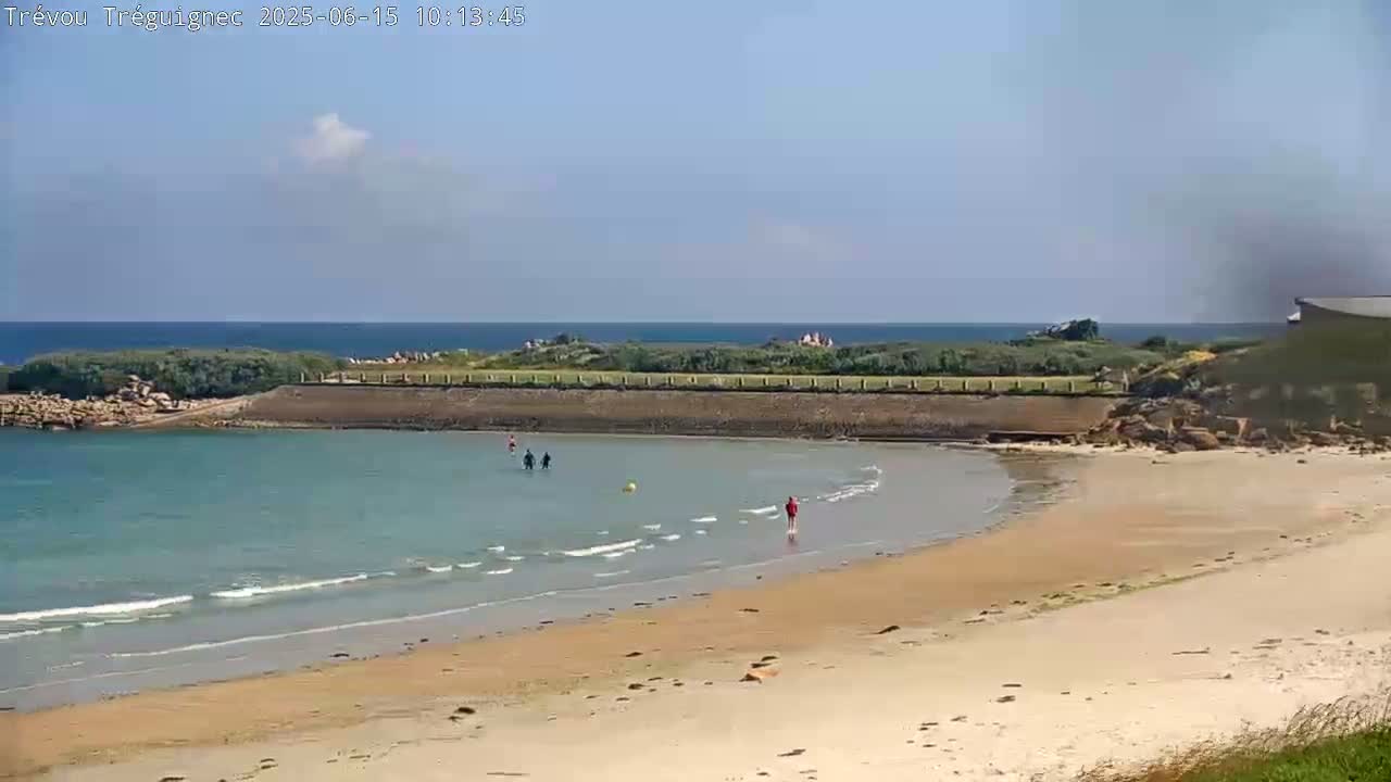 A sandy beach with calm, light-blue water laps gently at the shore under a mostly clear sky, with a few people wading in the water and walking along the beach.