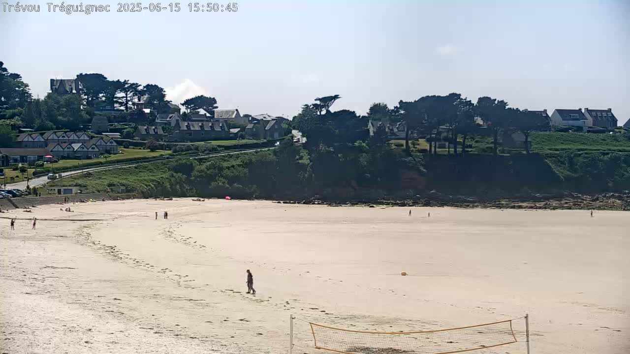 A sandy beach with a few people on it stretches below a small town nestled on a green hillside under a bright, sunny sky.