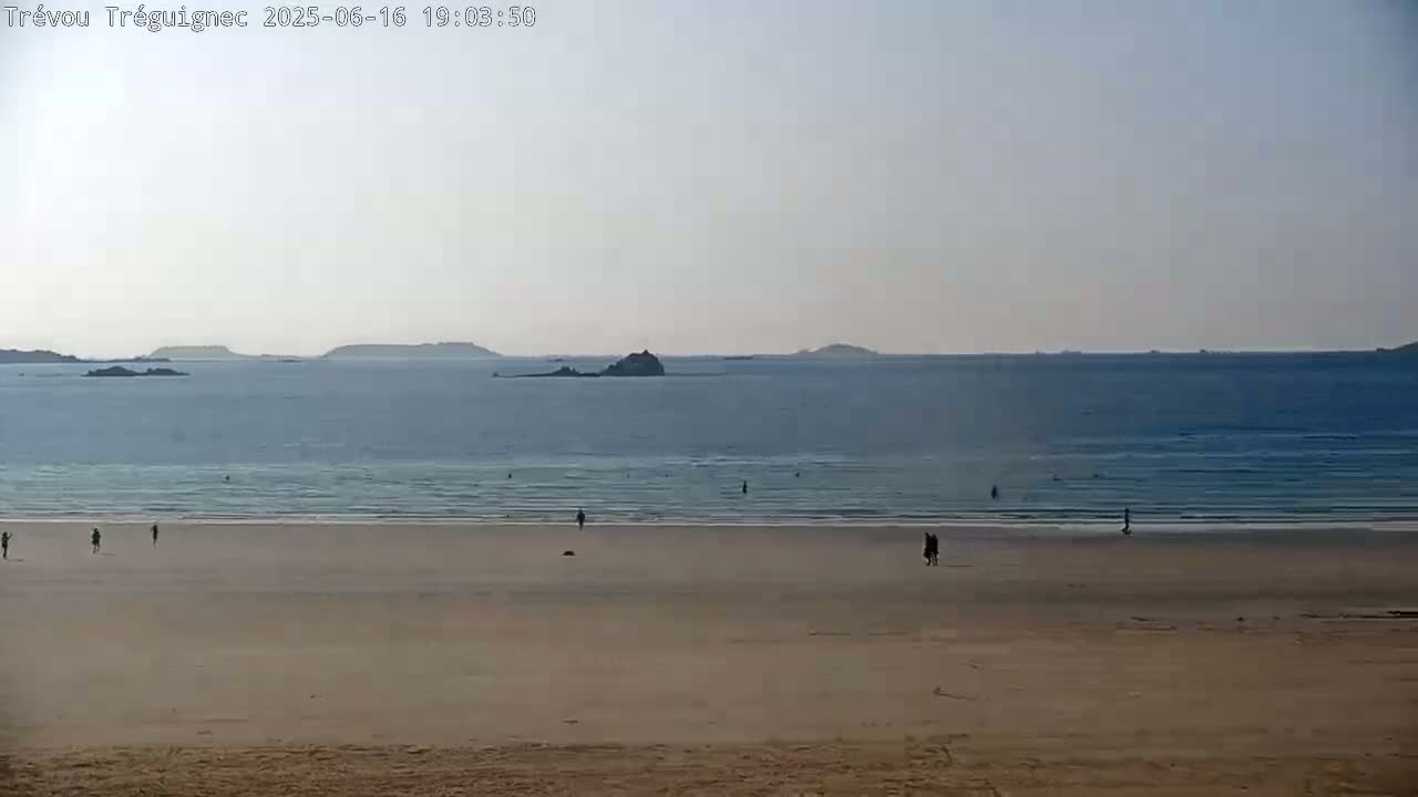 A sandy beach with a few people on it stretches to a calm ocean with distant islands under a clear, light sky.