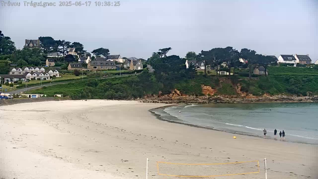 A sandy beach with gentle waves laps the shore, a few people walk along the water's edge, and houses are situated on a hill overlooking the beach under a partly cloudy sky.