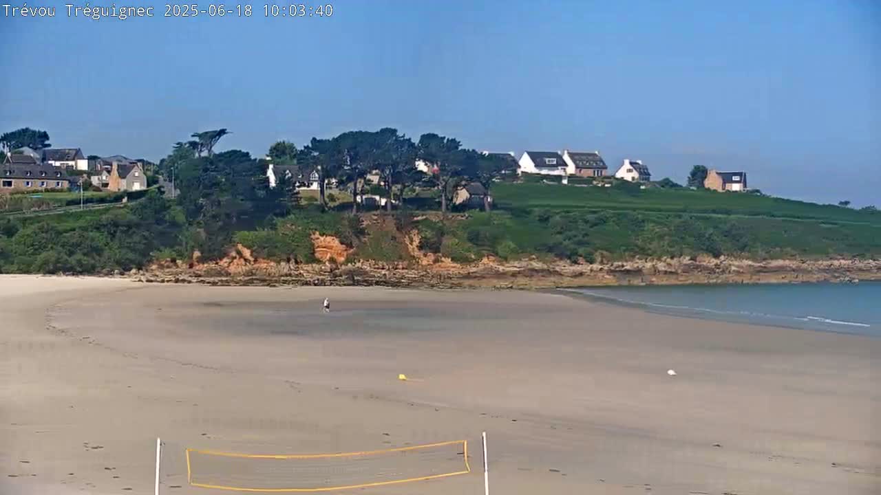A sandy beach with a volleyball net and a few people is situated below a grassy hillside with houses, under a clear blue sky.