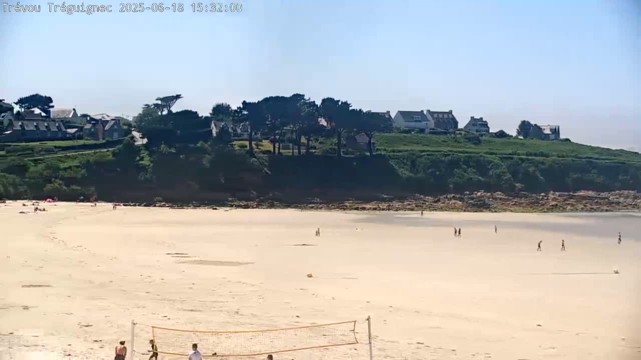 A sandy beach with a few people on it stretches out below a line of houses and green hillside on a sunny day.