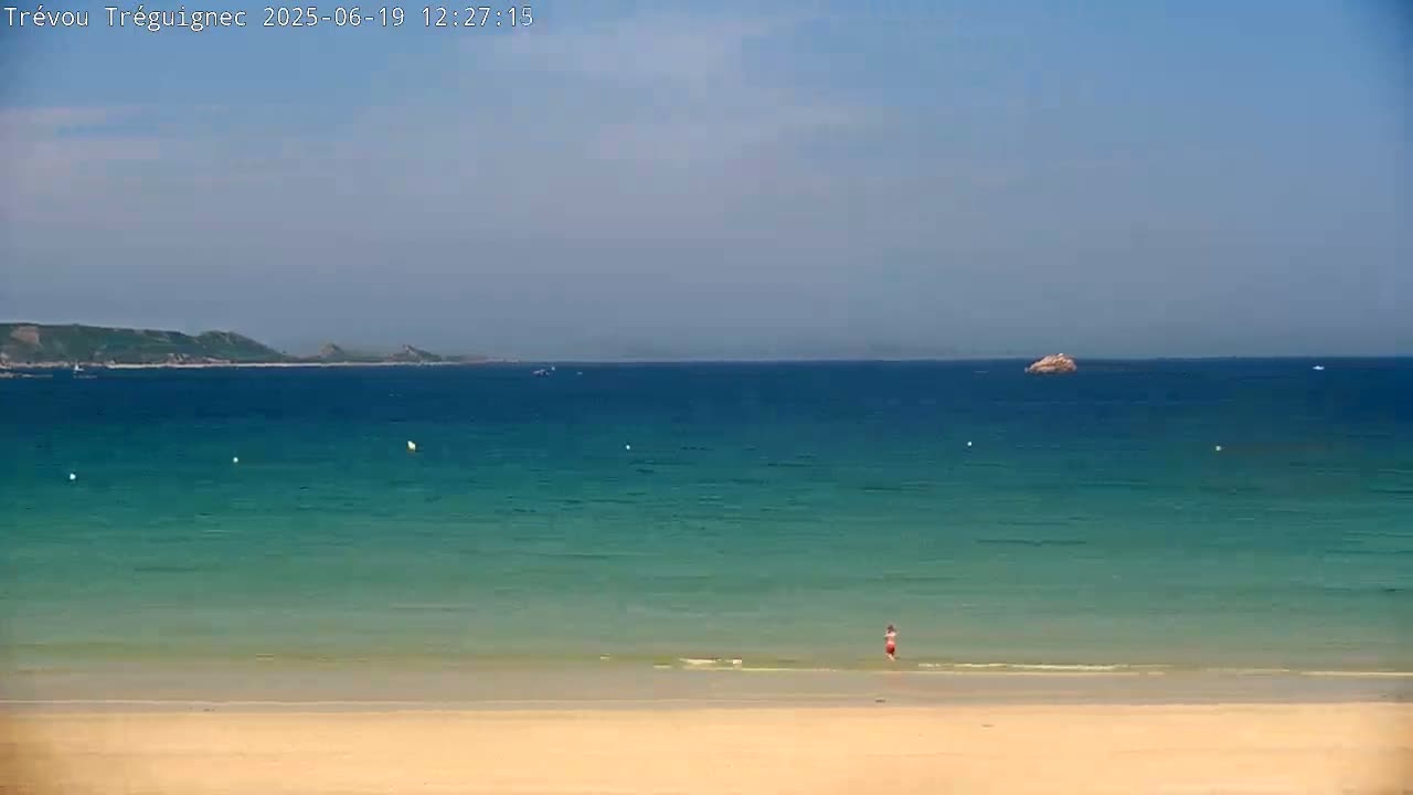 A person wades in shallow, clear turquoise water near a sandy beach, with a calm ocean and distant landmass visible under a mostly clear sky.
