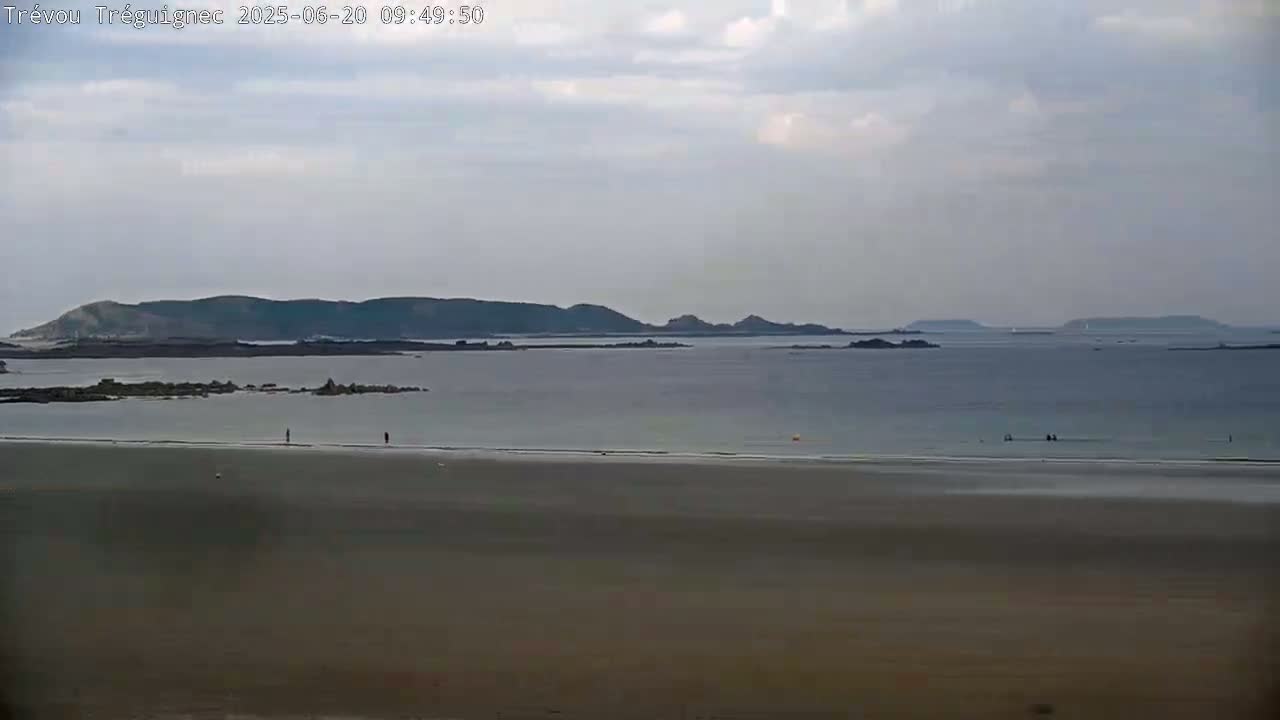 A long, flat sandy beach stretches to a calm, grey-blue ocean where a few people are visible in the water, with dark hills and islands visible in the distance under a mostly cloudy sky.