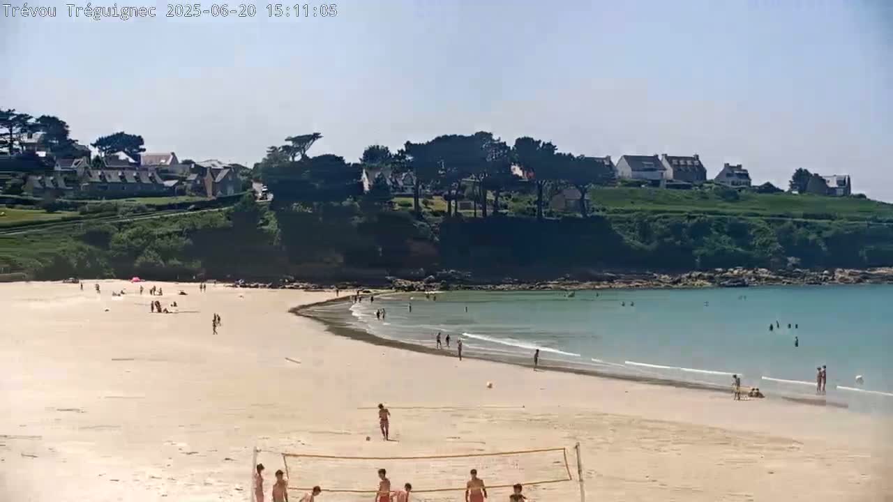 A sandy beach with calm, light-blue water and scattered people is backed by a green hillside with houses under a clear, sunny sky.