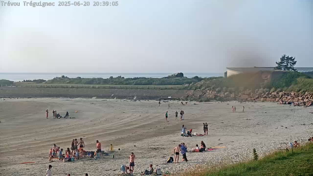 A sandy beach with a calm ocean in the background, under a hazy sky, is sparsely populated with people enjoying the late afternoon.