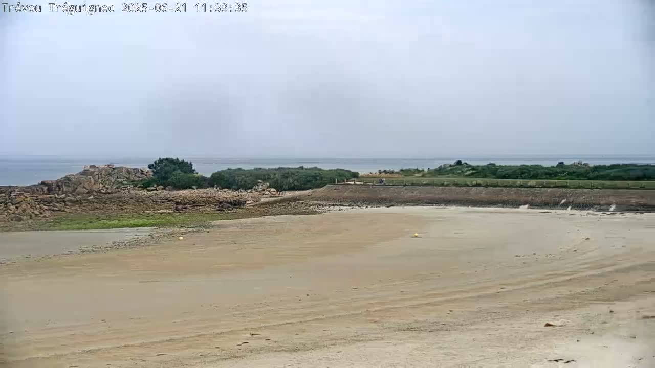 A sandy beach stretches to a rocky outcrop and calm ocean under an overcast sky.