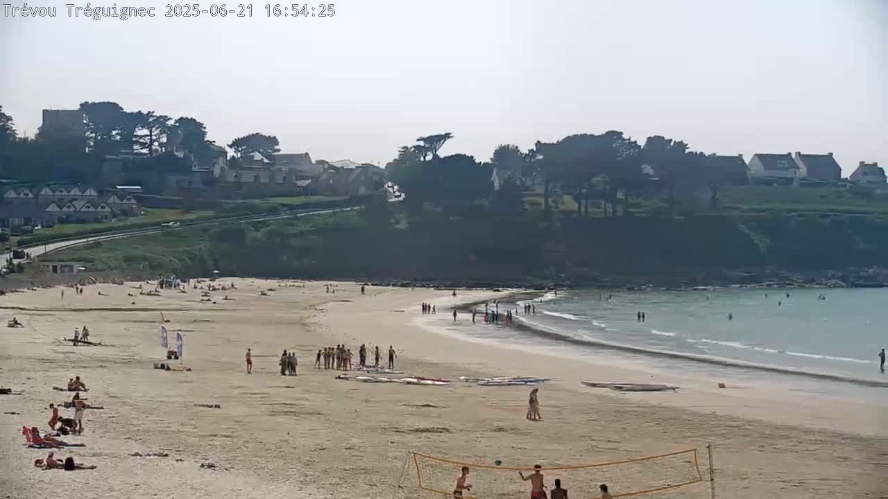 A sandy beach with numerous people sunbathing and swimming, bordered by a low cliff with houses and trees, on a hazy day.