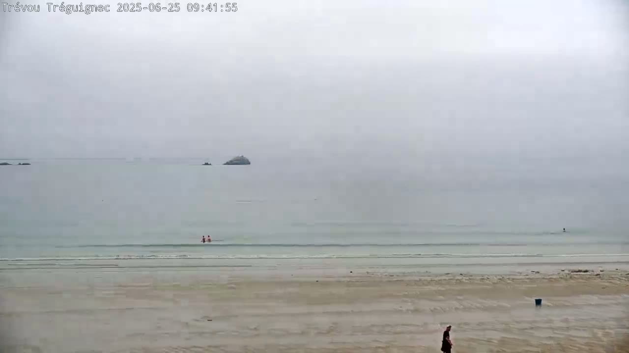 A mostly overcast day at a beach shows a person walking on the sand near calm, shallow water where a few people are swimming near small rocks visible in the distance.
