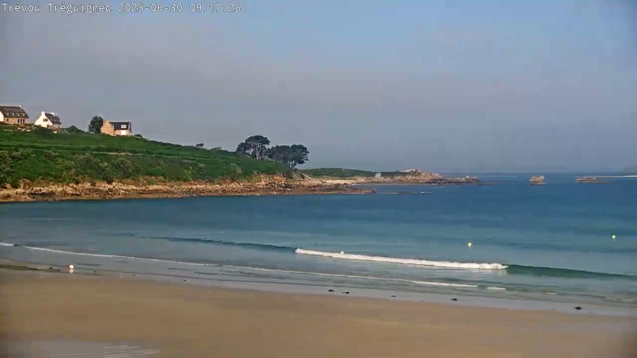 A sandy beach with small waves laps a calm bay, beyond which lies a rocky coastline with a few houses perched on a grassy hill under a clear sky.
